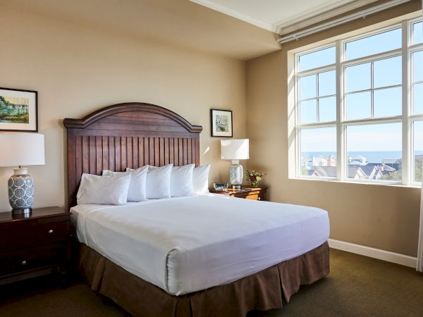 A cozy bedroom with a large bed, two lamps, framed art, and a window showcasing a view of the ocean and rooftops.