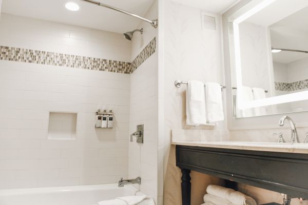 The image shows a modern bathroom with a bathtub, shower, tiled wall, towels, toiletries, and an illuminated mirror above the sink.