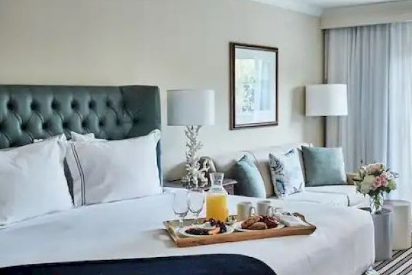 A cozy hotel room with a neatly made bed, breakfast tray, a sofa, and decor including a lamp and flower vase near a window.