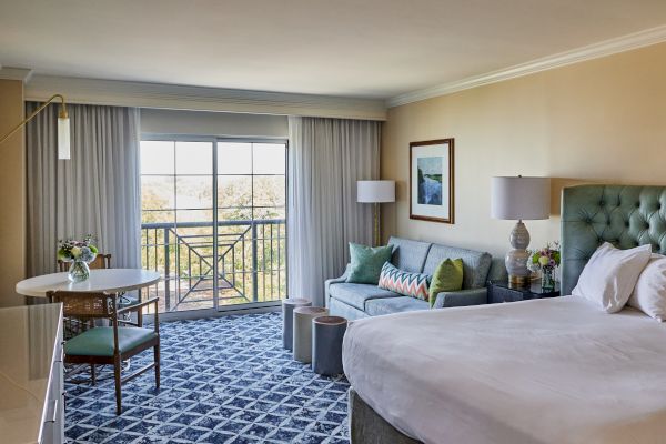 A well-lit hotel room with a bed, sofa, table, and chairs, featuring a balcony and decorative elements above the bed.