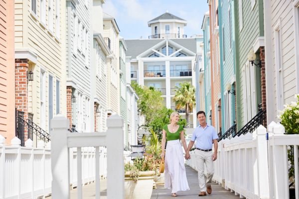 A couple holding hands, walking down a charming street lined with colorful buildings and white fences, on a sunny day.