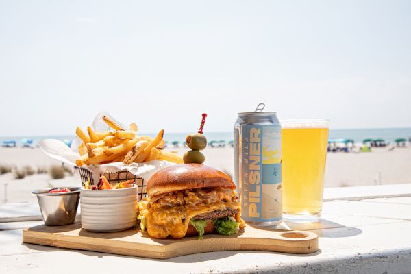 A burger with fries, sauces, a can of pilsner, and a glass are on a board by the beach with the ocean in the background.