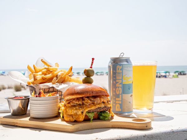 A burger with fries, sauces, a can of pilsner, and a glass are on a board by the beach with the ocean in the background.