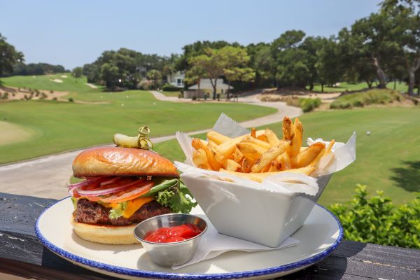 A delicious burger with fries and ketchup is served on a plate. The background shows a scenic golf course under a clear sky.