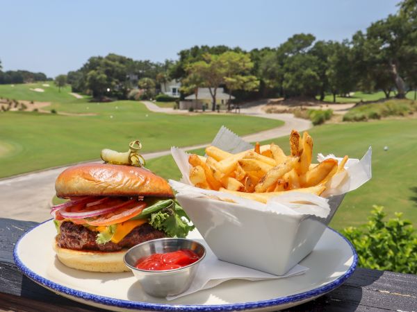 A delicious burger with fries and ketchup is served on a plate. The background shows a scenic golf course under a clear sky.