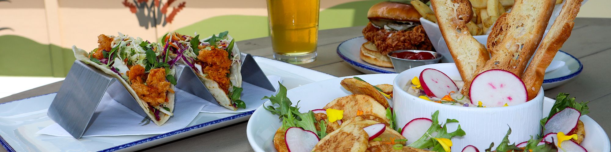 The image shows a variety of food including tacos, a sandwich, and a salad with a glass of beer on a table.