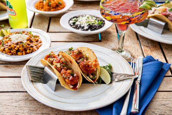 Two tacos on a plate with lime, alongside rice, beans, and a colorful beverage on a wooden table.