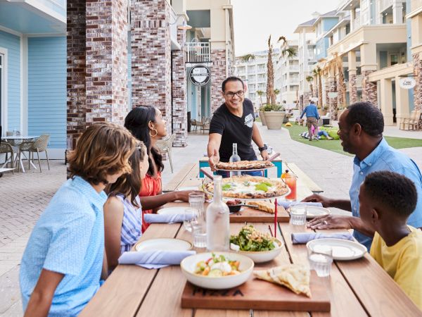 A group of people sitting at an outdoor table are being served pizza by a waiter.