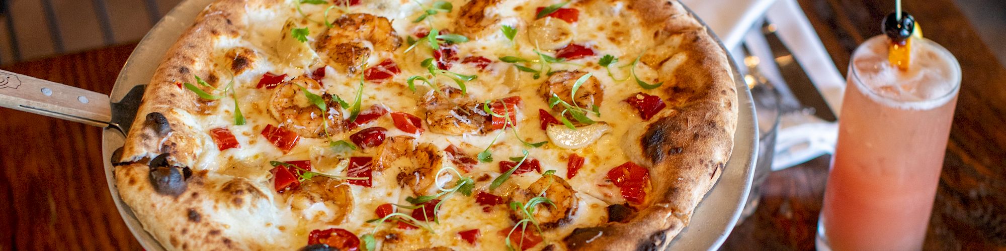 A wood-fired pizza, a salad, a dish with bread and cheese, and two drinks on a wooden table.