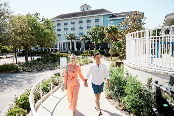 A couple walks hand in hand on a sunny path, with a multi-story building and greenery in the background.