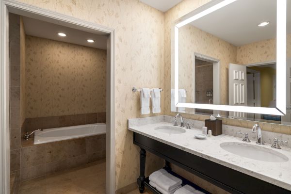 This image shows a luxurious bathroom with a double vanity, illuminated mirror, towels, and a spacious bathtub under recessed lighting.