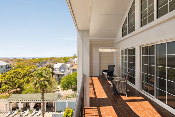 A balcony with chairs overlooks a scenic neighborhood with trees, houses, and a pool area under a clear blue sky.