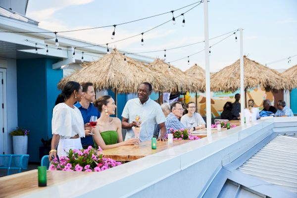 People enjoying drinks and conversation on a rooftop bar with string lights and thatched umbrellas in the background.