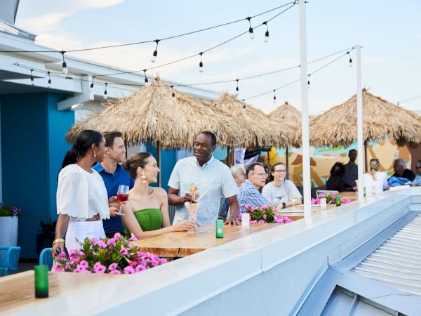 People enjoying drinks and conversation on a rooftop bar with string lights and thatched umbrellas in the background.