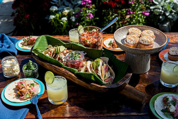 A table set with tacos, muffins, a jar of salsa, drinks with lime, and vibrant tableware, surrounded by flowers in the background.