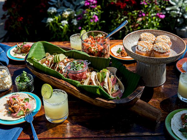 A table set with tacos, muffins, a jar of salsa, drinks with lime, and vibrant tableware, surrounded by flowers in the background.