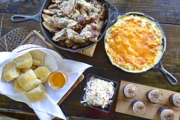 A spread with pulled pork, mac and cheese, rolls with sauce, coleslaw, and chocolate cupcakes.