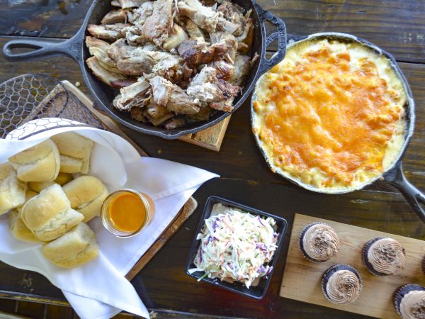 A spread with pulled pork, mac and cheese, rolls with sauce, coleslaw, and chocolate cupcakes.
