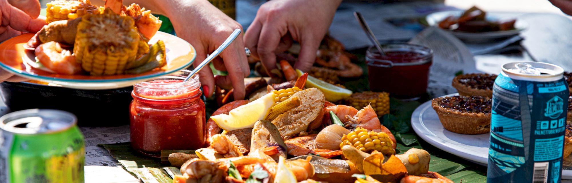 A seafood boil with corn, shrimp, and dipping sauces, surrounded by drinks and pies, being served on a table outdoors.