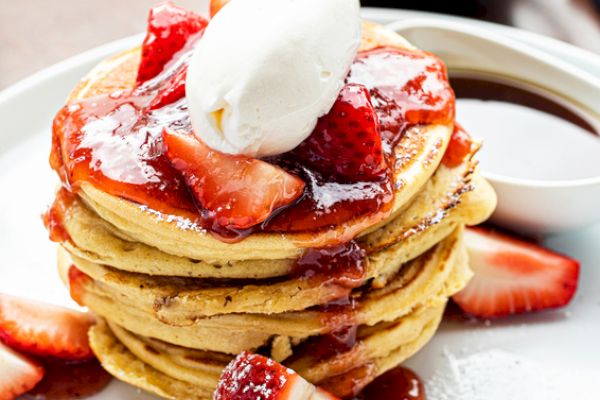 A stack of pancakes topped with strawberries, syrup, and a dollop of cream, served on a white plate with additional strawberry slices.