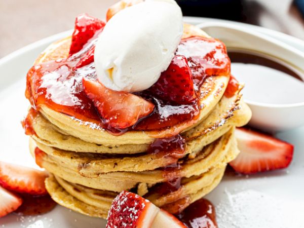 A stack of pancakes topped with strawberries, syrup, and a dollop of cream, served on a white plate with additional strawberry slices.