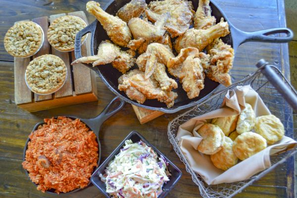 The image shows fried chicken, coleslaw, rice, biscuits in a basket, and baked dishes on a wooden table.