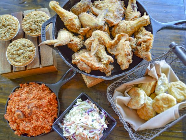The image shows fried chicken, coleslaw, rice, biscuits in a basket, and baked dishes on a wooden table.
