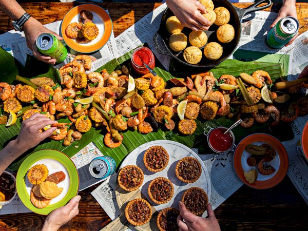 An overhead view of a seafood boil spread with crab cakes, corn, and assorted drinks on a table with people reaching for food.