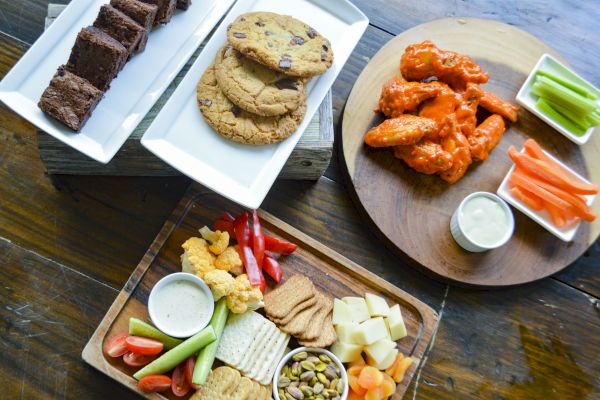 The image shows a spread of desserts, cookies, buffalo wings with dip, and a platter of veggies, cheese, crackers, and snacks on a table.