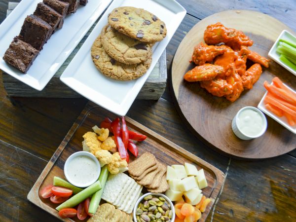 The image shows a spread of desserts, cookies, buffalo wings with dip, and a platter of veggies, cheese, crackers, and snacks on a table.