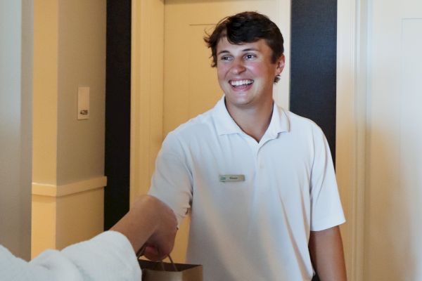 A person in a white shirt is smiling and receiving a bag from another person in a hallway.