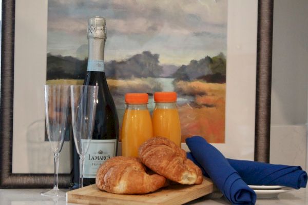 A bottle of sparkling wine, two juice bottles, croissants, flutes, and a blue napkin on a board, against a scenic painting backdrop.