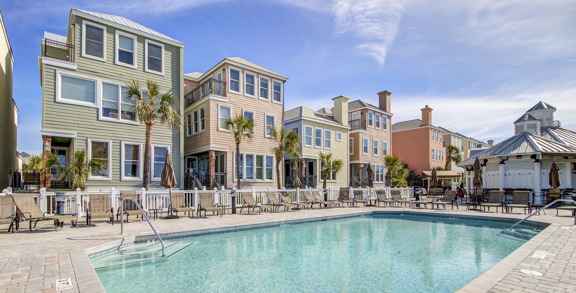 A swimming pool surrounded by colorful buildings and palm trees, with lounge chairs arranged neatly on a sunny day under a blue sky.