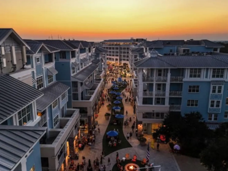 A lively street scene at sunset features modern buildings, a crowd of people, outdoor seating, and a decorative fire pit in the center.