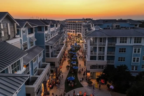 A lively street scene at sunset features modern buildings, a crowd of people, outdoor seating, and a decorative fire pit in the center.