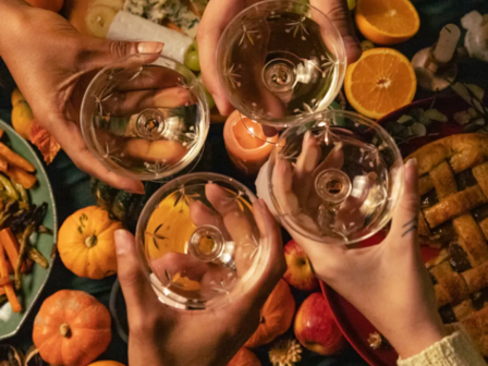 People are holding glasses for a toast over a table filled with pumpkins, fruits, and various foods, creating a festive atmosphere.
