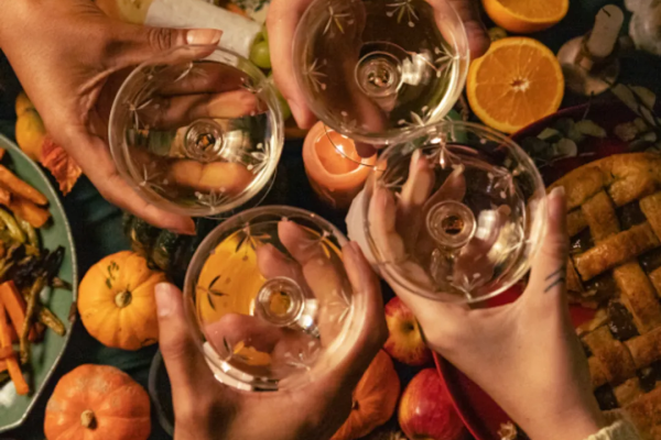 People are holding glasses for a toast over a table filled with pumpkins, fruits, and various foods, creating a festive atmosphere.