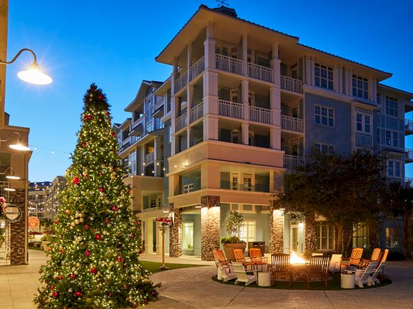 A decorated Christmas tree stands near a residential building at night, surrounded by a lit seating area and soft evening lights.