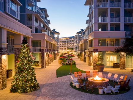 An outdoor plaza with a Christmas tree, surrounded by buildings, chairs around a lit fire pit, and evening lighting.