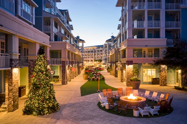 An outdoor plaza with a Christmas tree, surrounded by buildings, chairs around a lit fire pit, and evening lighting.