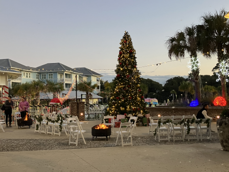 An outdoor holiday setup with a Christmas tree, string lights, chairs, and festive decorations, including palm trees.