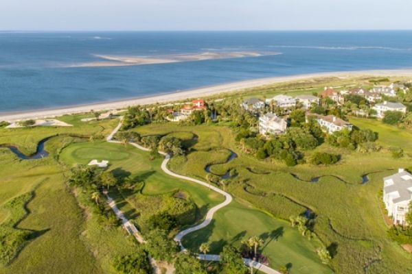 Aerial view of a coastal area with a golf course, winding paths, houses, and the ocean in the background.