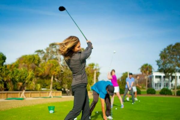 People are practicing golf on a lush, sunny course. A woman swings a club, while others prepare their shots in the background.