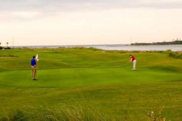Two people are playing golf on a green golf course with a water body in the background under a cloudy sky.