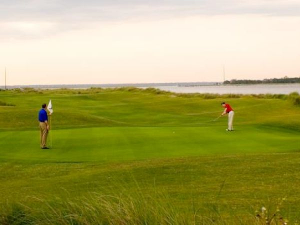 Two people are playing golf on a green golf course with a water body in the background under a cloudy sky.
