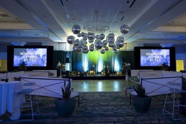 The image shows an event space with a stage, large screens, and decorative hanging spheres, titled "Midnight at the Dunes."