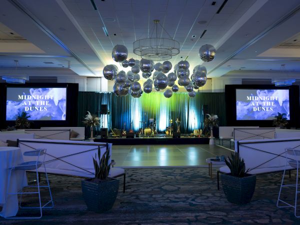 The image shows an event space with a stage, large screens, and decorative hanging spheres, titled "Midnight at the Dunes."