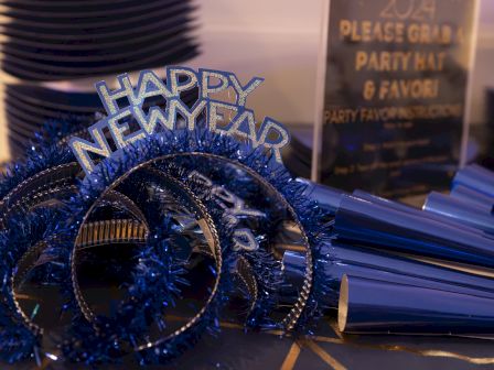 The image shows blue "Happy New Year" headbands and party hats on a table, with a sign inviting guests to grab them.