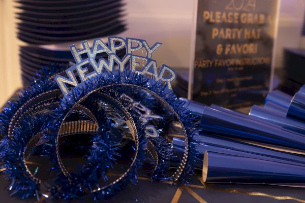 The image shows blue "Happy New Year" headbands and party hats on a table, with a sign inviting guests to grab them.