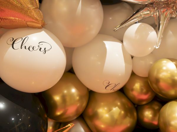 The image shows a festive arrangement of white, gold, and black balloons, with some displaying the word "Cheers."